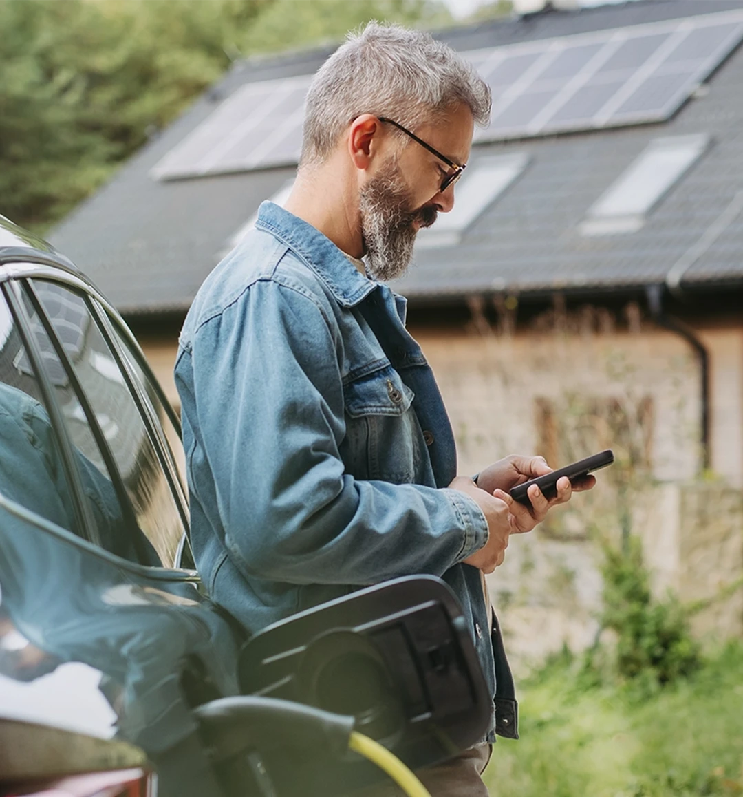 Mann lädt sein Elektroauto vor einem Haus mit Solaranlage auf dem Dach und steuert die Energieversorgung per Smartphone.