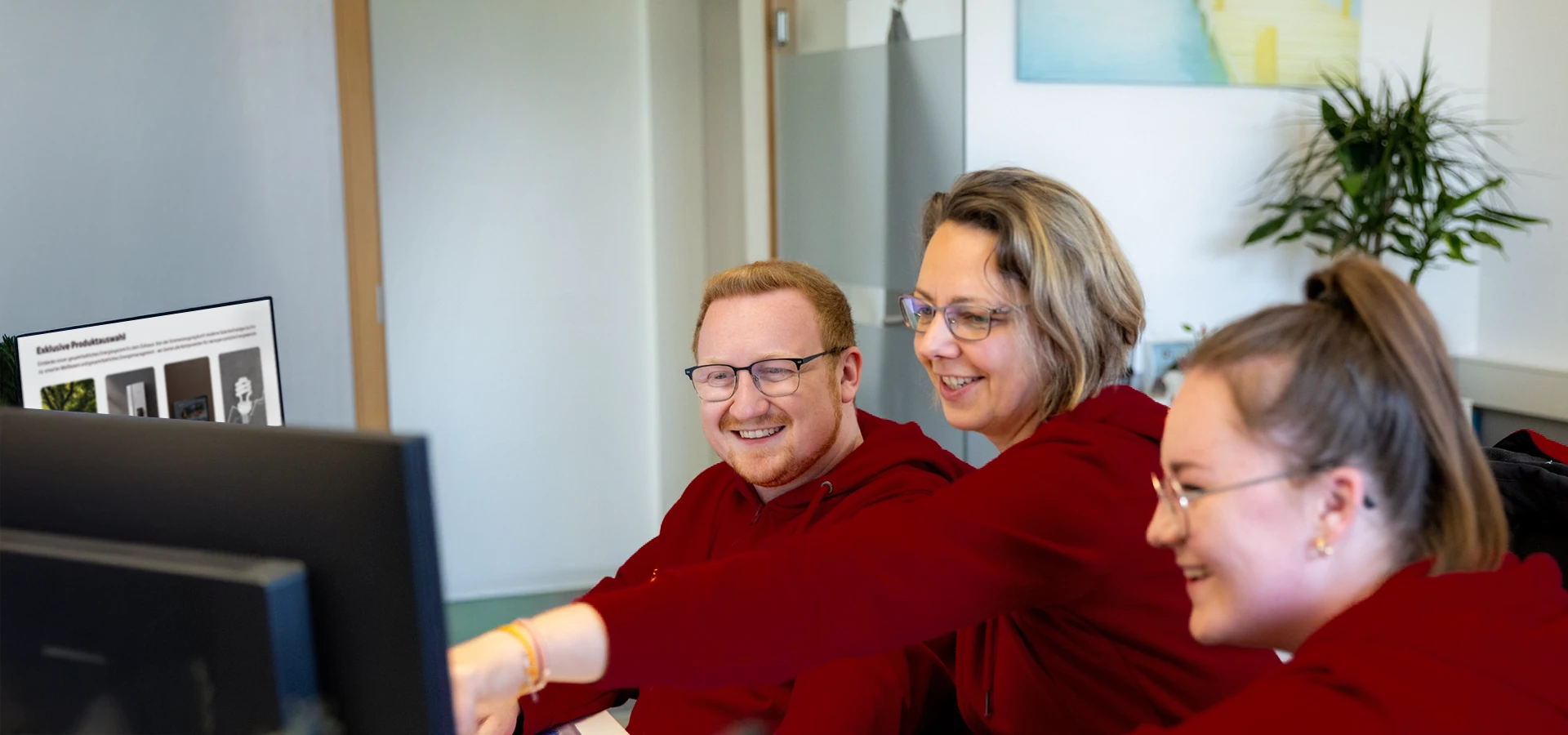 Drei BSH Mitarbeitende in roten Oberteilen sitzen gemeinsam vor einem Computerbildschirm und lachen, während eine Person auf den Monitor zeigt.