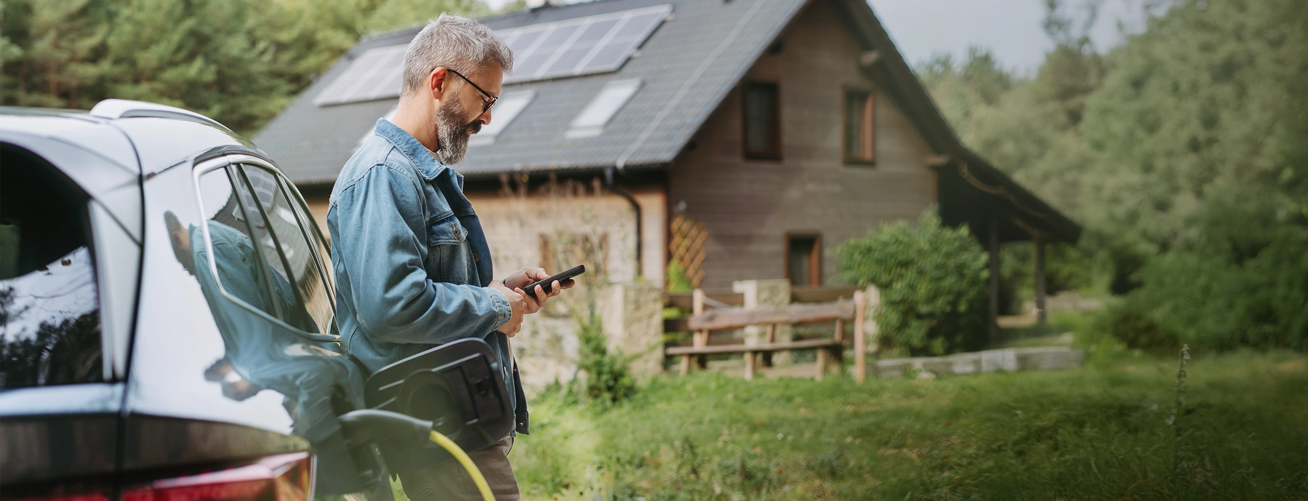Mann lädt sein Elektroauto vor einem Haus mit Solaranlage auf dem Dach und steuert die Energieversorgung per Smartphone.