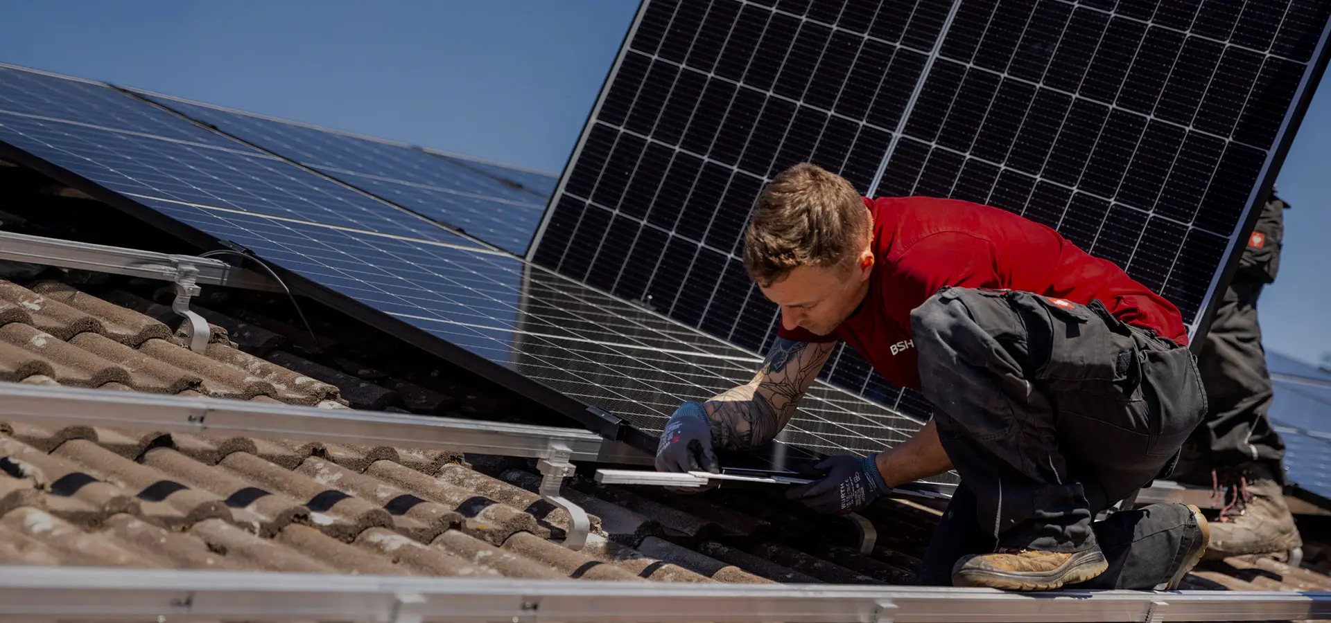 BSH Techniker in rotem Shirt montiert eine Photovoltaikanlage auf einem Ziegeldach bei Sonnenschein.