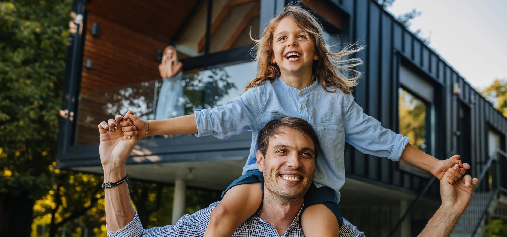 Vater trägt seine lachende Tochter auf den Schultern vor einem modernen Haus, im Hintergrund winkt eine Frau vom Balkon.