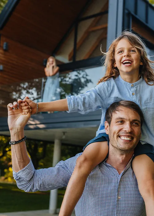 Vater trägt seine lachende Tochter auf den Schultern vor einem modernen Haus, im Hintergrund winkt eine Frau vom Balkon.