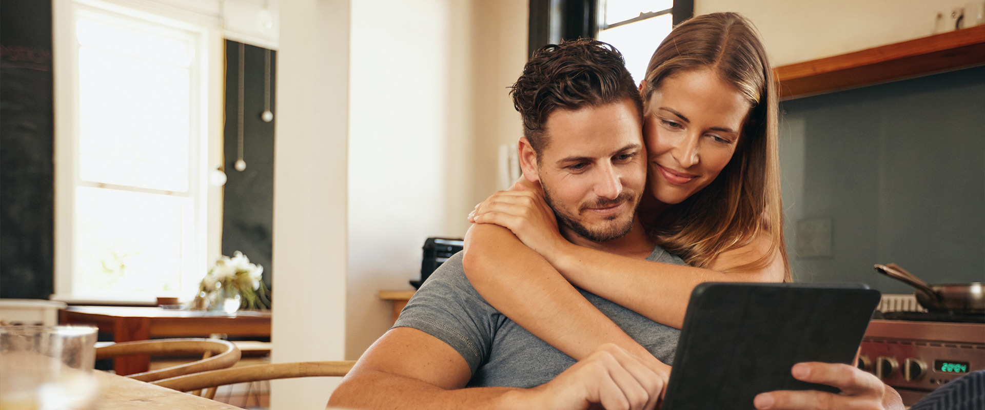 Ein Paar sitzt in einer hellen Küche, die Frau legt dem Mann die Arme um die Schultern, während beide gemeinsam auf ein Tablet schauen.
