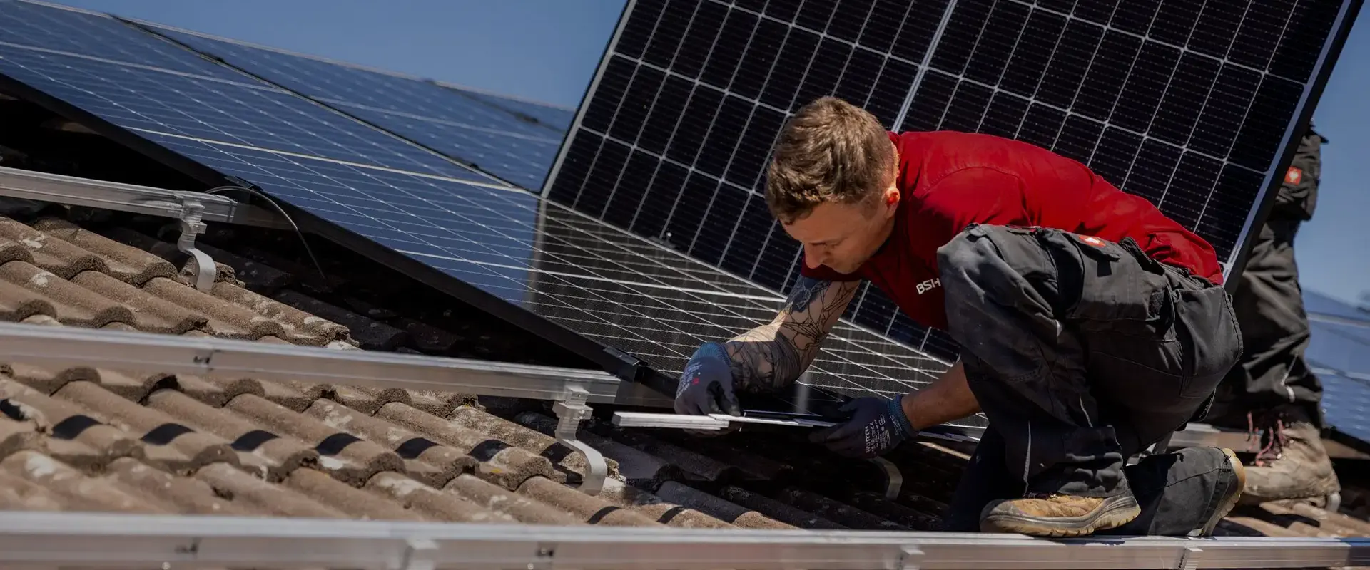 BSH Techniker in rotem Shirt montiert eine Photovoltaikanlage auf einem Ziegeldach bei Sonnenschein.