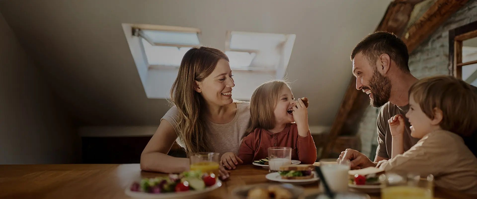 Lächelnde Familie mit zwei kleinen Kindern sitzt gemeinsam am Esstisch unter dem Dach und genießt eine Mahlzeit.