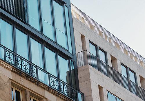 Moderne Gebäudefassade mit klaren Linien, großen Glasflächen und Balkonen, kombiniert mit einer hellen Steinfassade. Mischung aus historischer Architektur und zeitgenössischem Design.