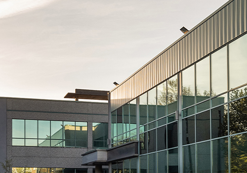 Moderne Büro- oder Gewerbeimmobilie mit großflächiger Glasfassade. In den Fenstern spiegeln sich Bäume und Himmel, daneben stehen begrünte Außenflächen. Aufnahme bei warmem Tageslicht.